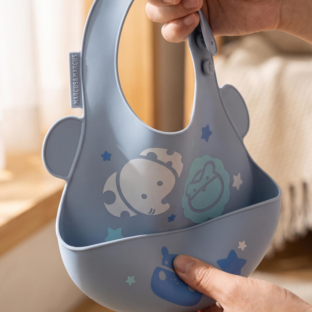 Marcus & Marcus pastel blue silicone baby bib with a handle, decorated with cute cartoon animal and star patterns, being held by hands in a softly lit indoor setting.