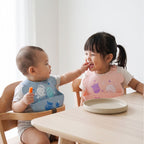  baby and a young girl sitting in wooden high chairs at a light wood table, wearing Marcus & Marcus silicone bibs. The baby wears a pastel blue bib with cartoon animals, and the girl wears a pastel pink bib with cartoon characters.