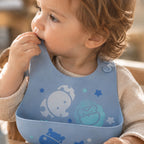 A close-up of a baby wearing Macus & Marcus pastel blue silicone bib with cartoon animal and star patterns, sitting in a high chair.