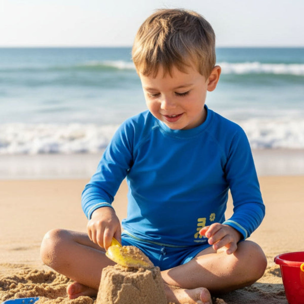 Child wearing long sleeve rash guard with marckids logo building a sandcastle on a beach with ocean in the background
