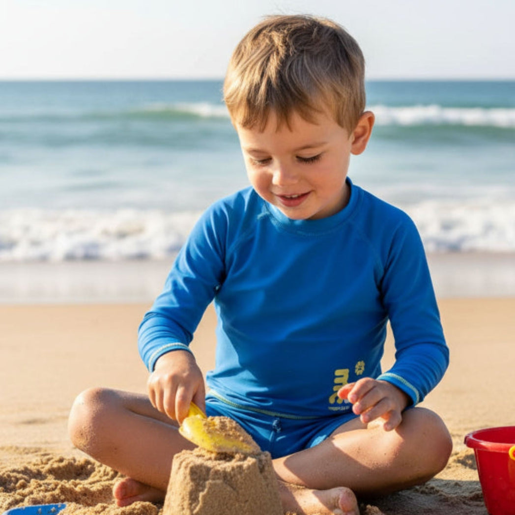 Child wearing long sleeve rash guard with marckids logo building a sandcastle on a beach with ocean in the background
