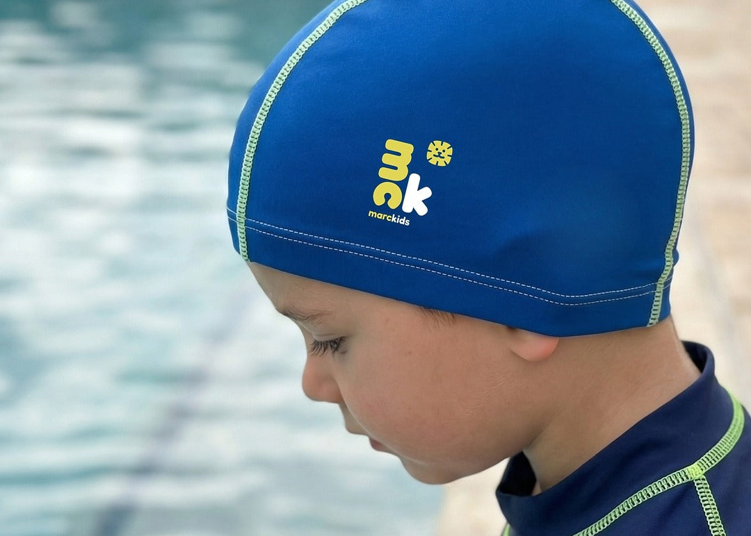 Child wearing marckids blue swim cap with a brand logo, standing by a pool.