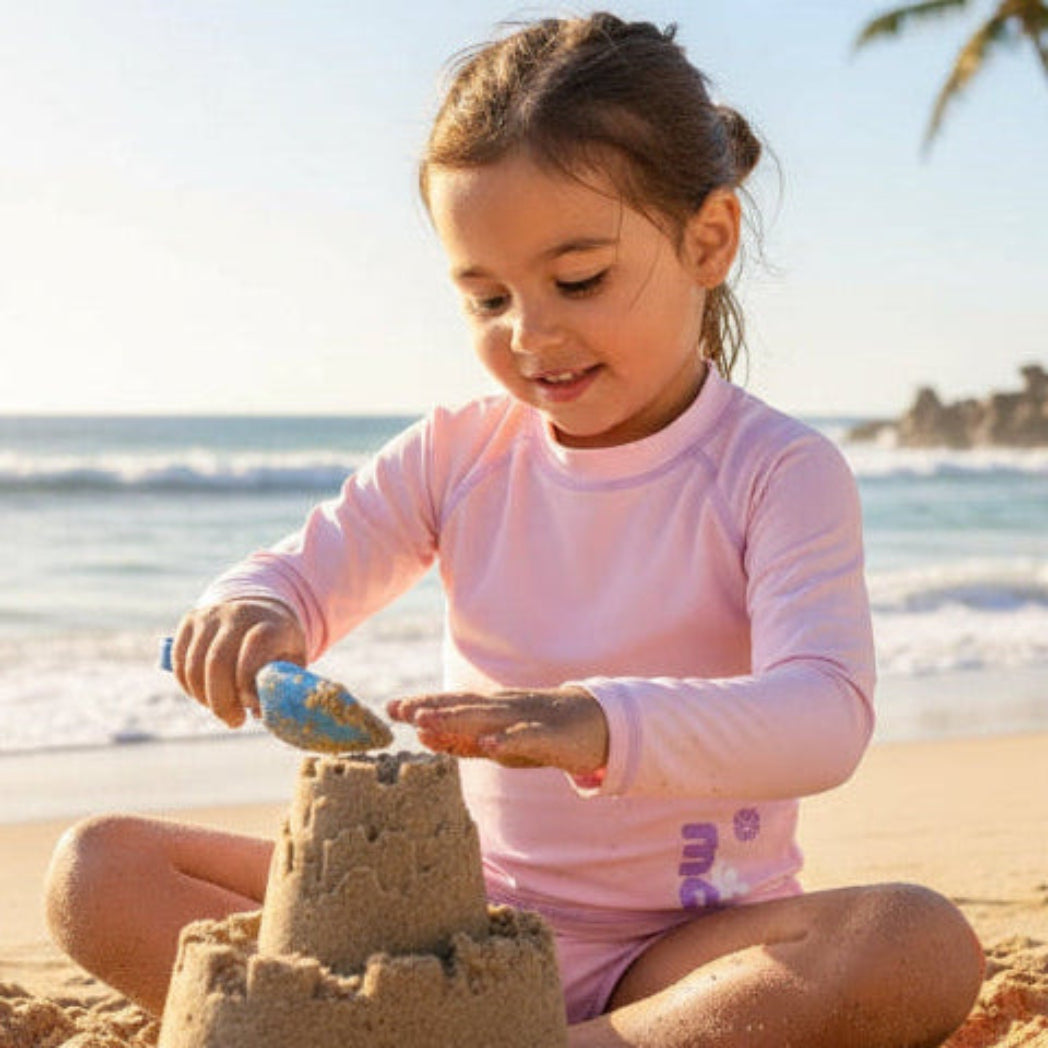 Child with long sleeve rash guard with marckids logo building a sandcastle on a beach with palm trees in the background